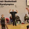 Wheelchair basketball players in action during a 2024 Paralympics game, with a referee and spectators in the background.